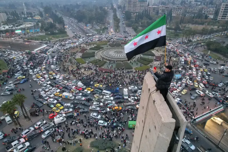 شخص يرفع علم سوريا فوق مبنى بإحدى ميادين سوريا